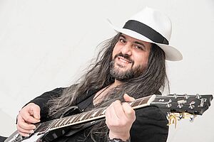 Raffaele E. Quarta mit weißem Hut und E-Gitarre vor weißem Hintergrund