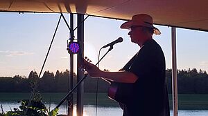 Sänger Robert Wildfeuer mit Mikrofon und Gitarre vor einem See und Wald 